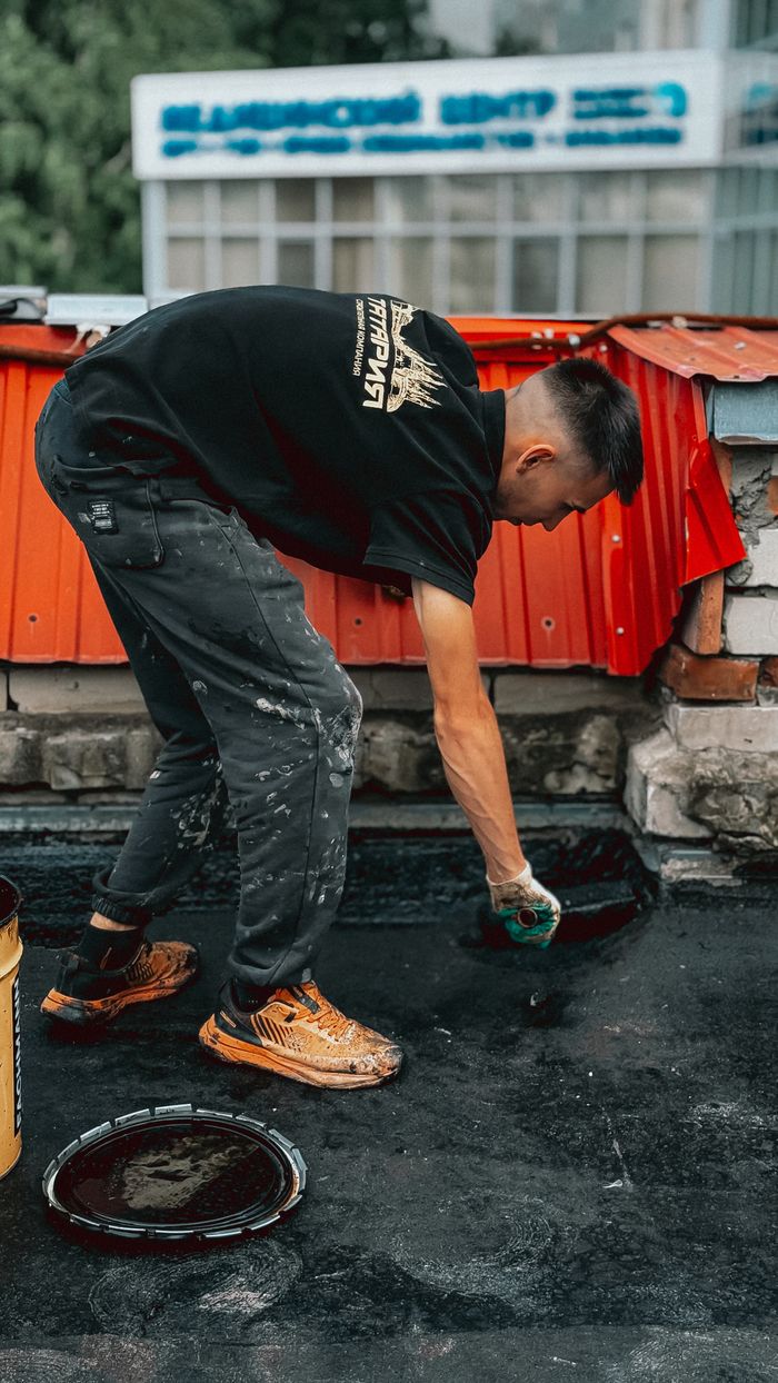 A young man is applying waterproofing material on a rooftop, wearing casual workwear.